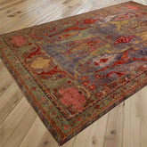 A rectangular area rug with an intricate traditional pattern in burnt orange, grey, soft green, gold and rose pink, shown laid out on a light wood plank floor.
