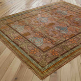 A rectangular area rug with an ornate floral and vine pattern, shown laid out on a light-colored wooden plank floor. The rug features a warm brown base with prominent orange, sage green, and light blue accents.