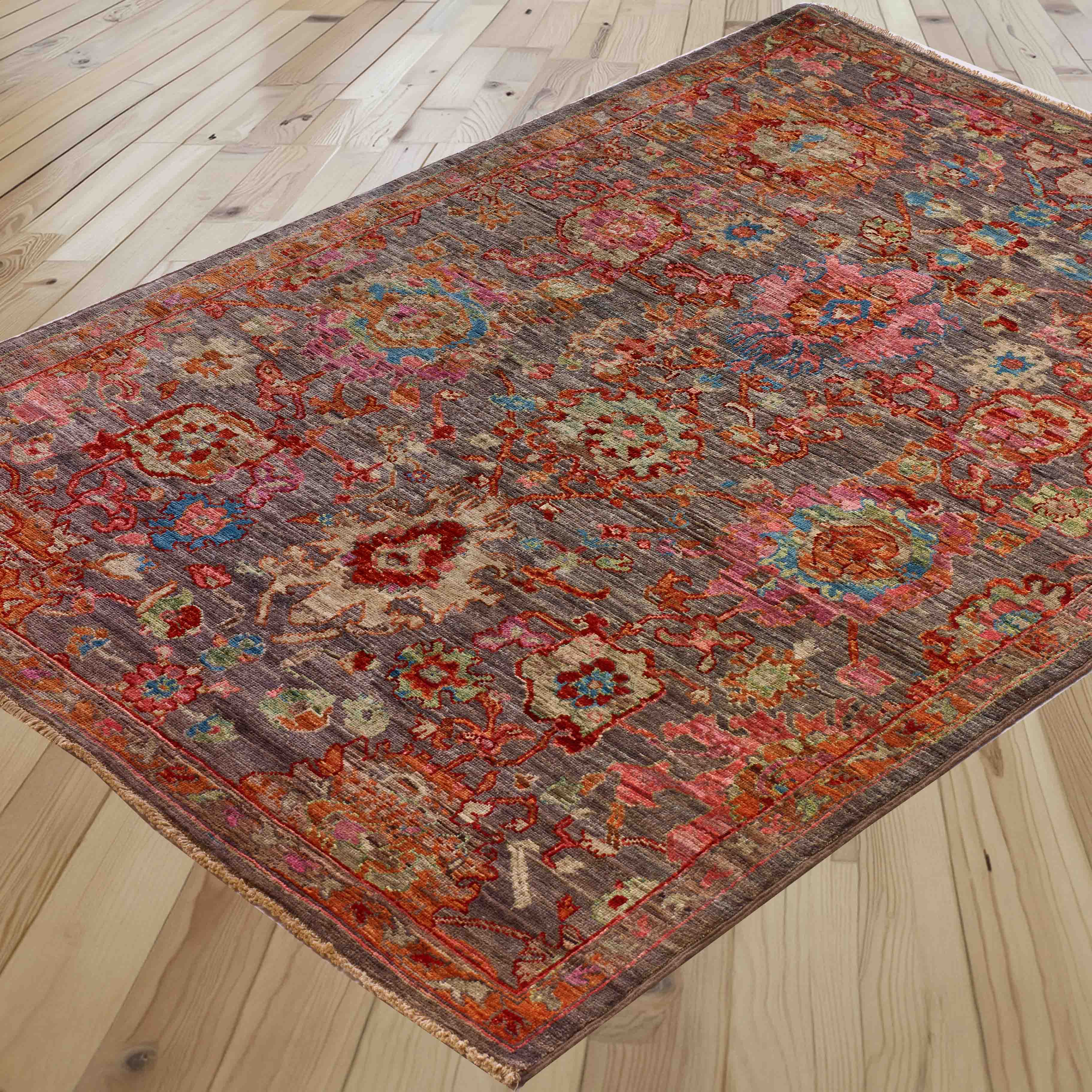 A rectangular area rug with a traditional floral pattern in sage green, rose pink, heather, and terracotta, shown laid out on a light-colored wooden floor.