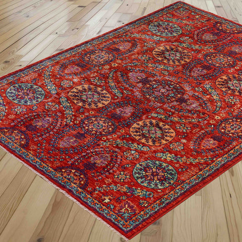 A traditional area rug with a vibrant red base and intricate multicolour floral patterns, shown laid out on a light wood plank floor.