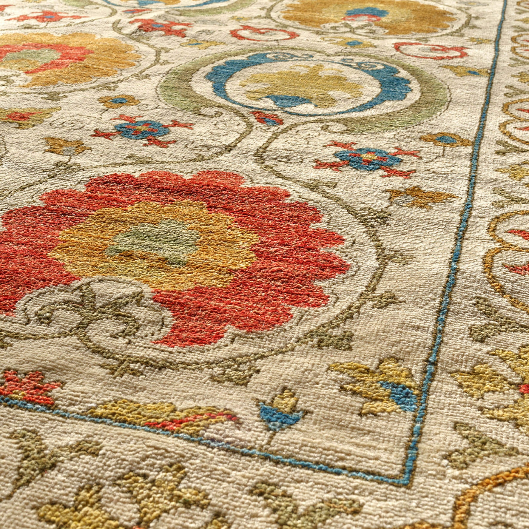 Close-up of a woven area rug with an ornate floral and scroll pattern in red, yellow, blue, and olive on a warm cream background.